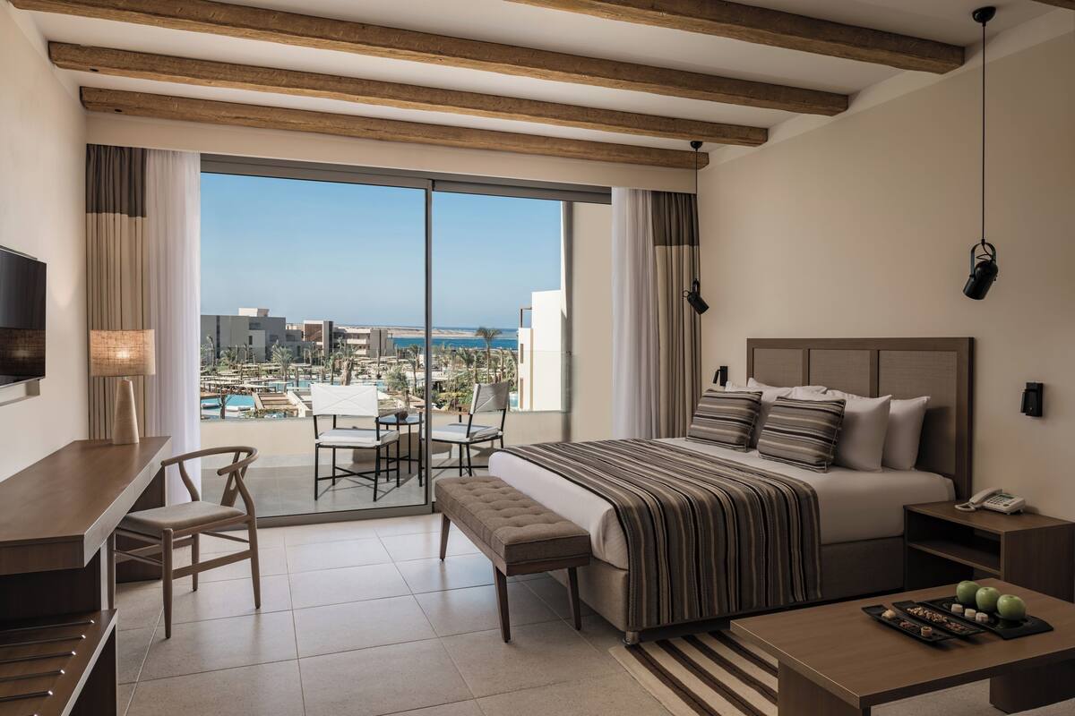 A hotel room with a large bed, a desk, a chair, a small table, a bench, a bedside table, and a view of the city and ocean.