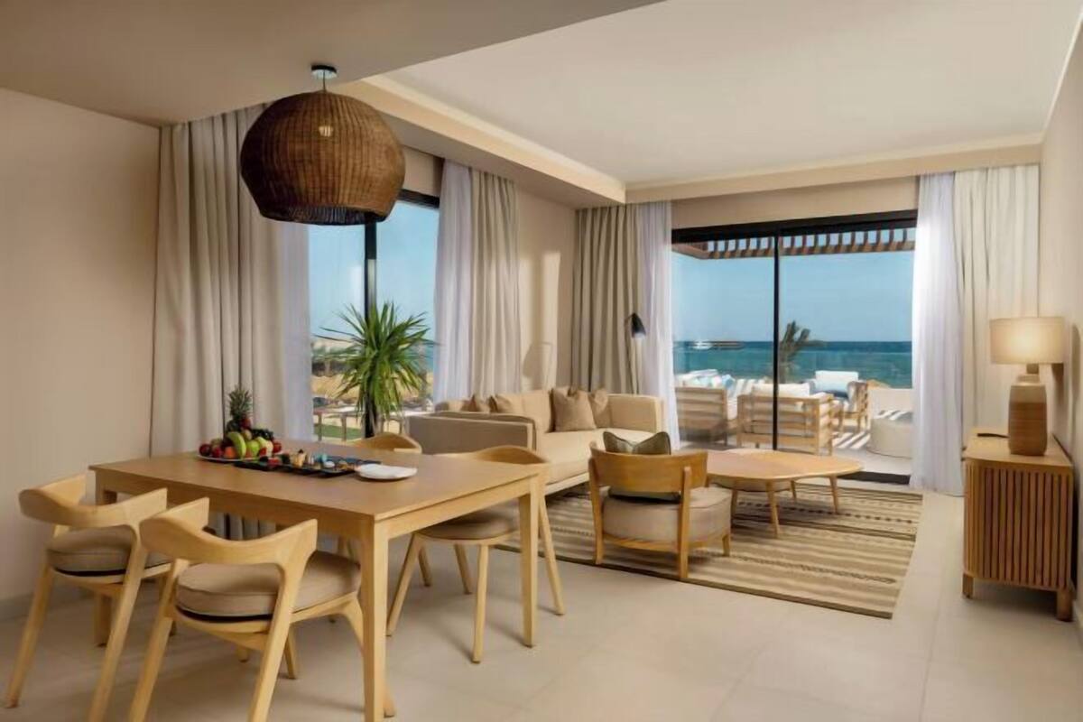 A modern hotel room with a dining area, a sofa set, a large window with a view of the sea, and a hanging light fixture.