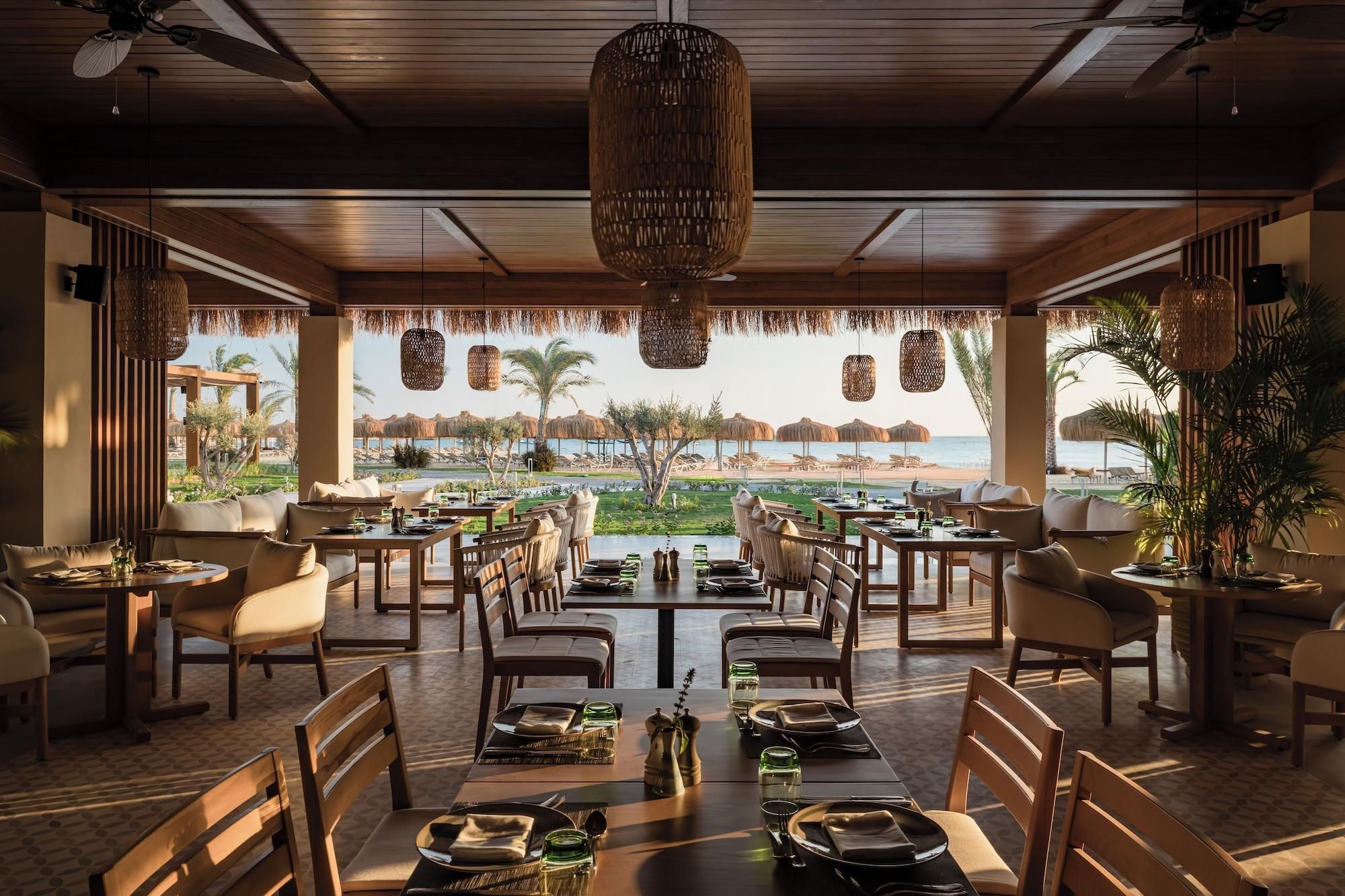 Un restaurante con mesas dispuestas para comer, vista a la playa y detalles decorativos en techo de paja.