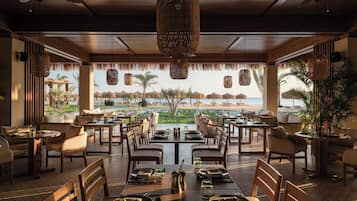 Un restaurante con mesas dispuestas para comer, vista a la playa y detalles decorativos en techo de paja.