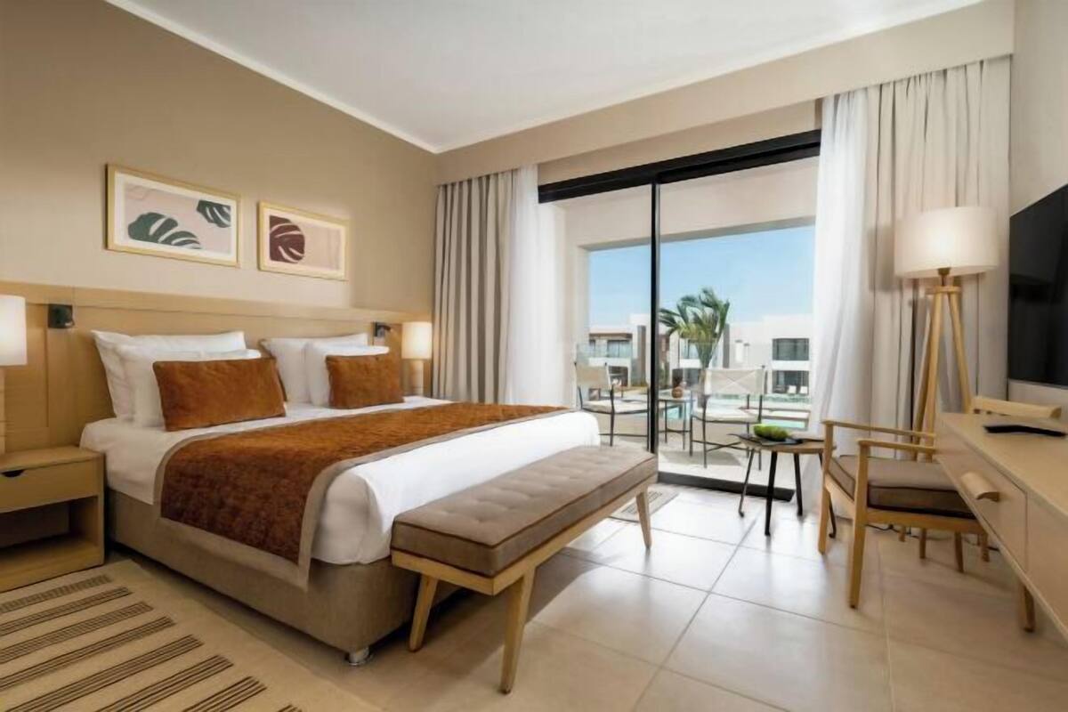 A hotel room with a bed, bedside tables, a desk, a chair, a TV, and a balcony with a view.