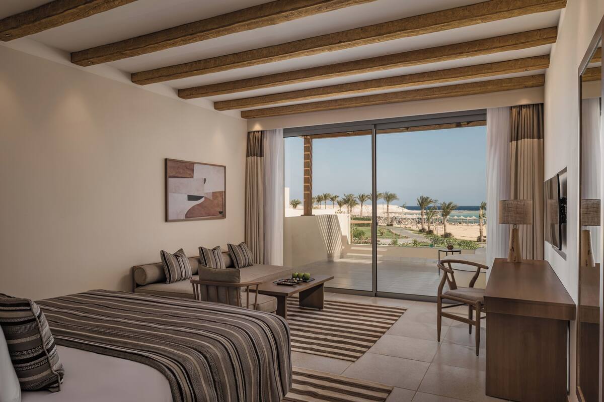 A hotel room with a bed, a sofa, a coffee table, a TV, and a balcony with a view of the beach.