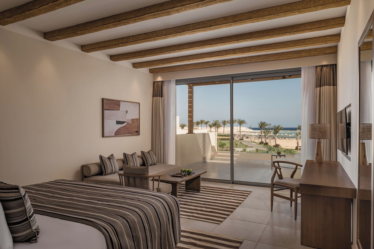 A hotel room with a bed, a sofa, a coffee table, a TV, and a balcony with a view of the beach.
