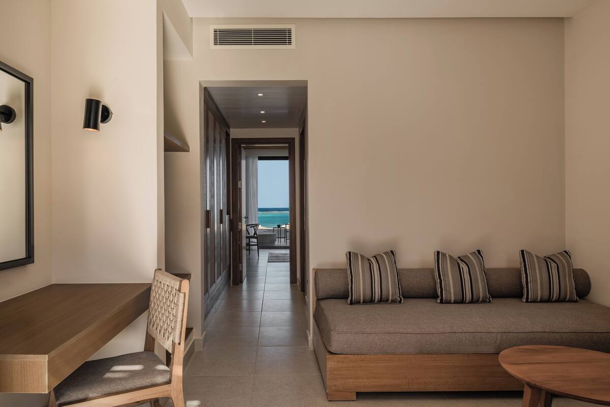 A modern hotel room with a wooden desk, a cushioned chair, a sofa with striped pillows, and a view of the ocean through an open doorway.