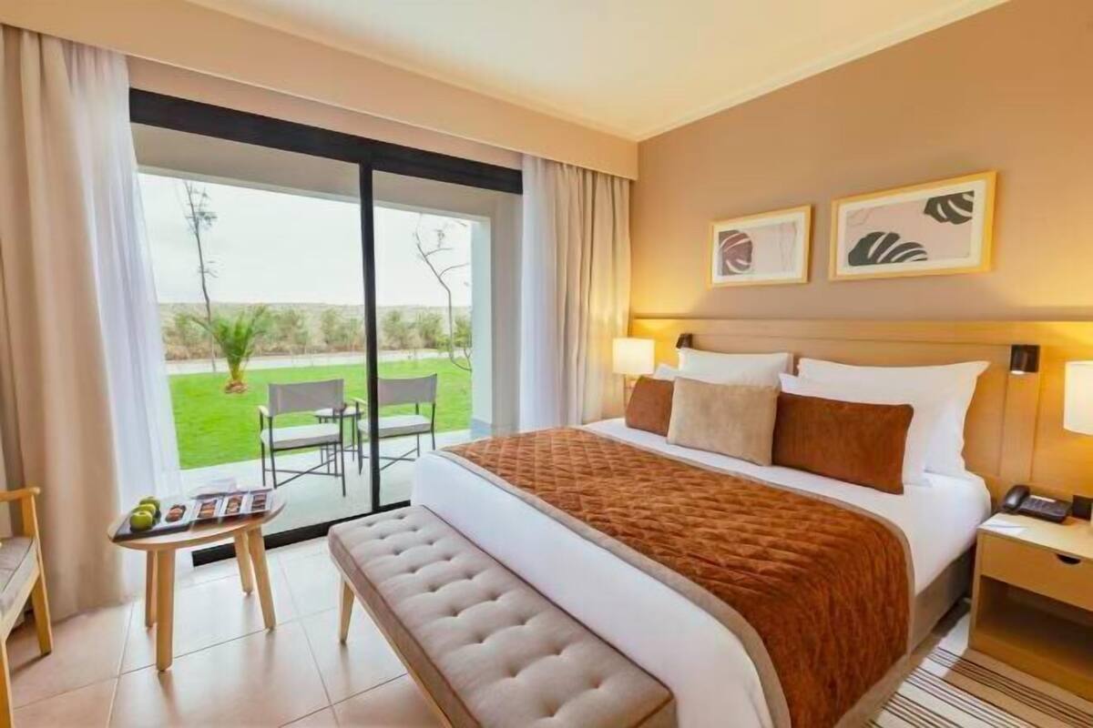 A hotel room with a large bed, a bench, a table with a tray of fruits, a chair, and a view of a green landscape through a sliding door.