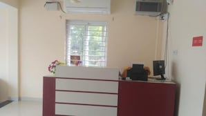 A reception desk with a computer, a small plant, and a window with blinds. - MM Park Inn (Mahabalipuram)