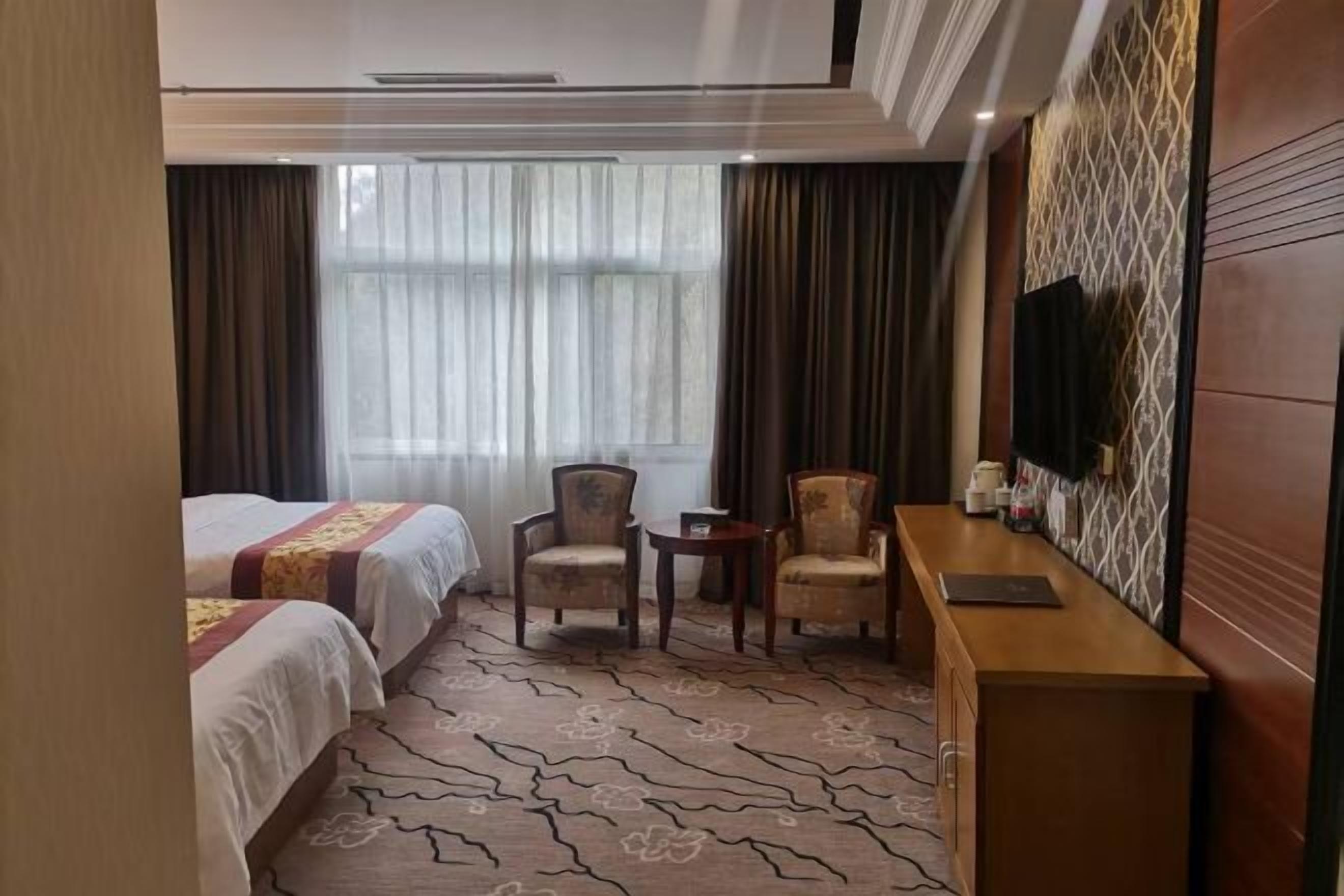 Hotellihuone, jossa on kaksi sänkyä, pieni oleskelualue tuoleineen, puinen sivupöytä, televisio ja kuviollinen matto.