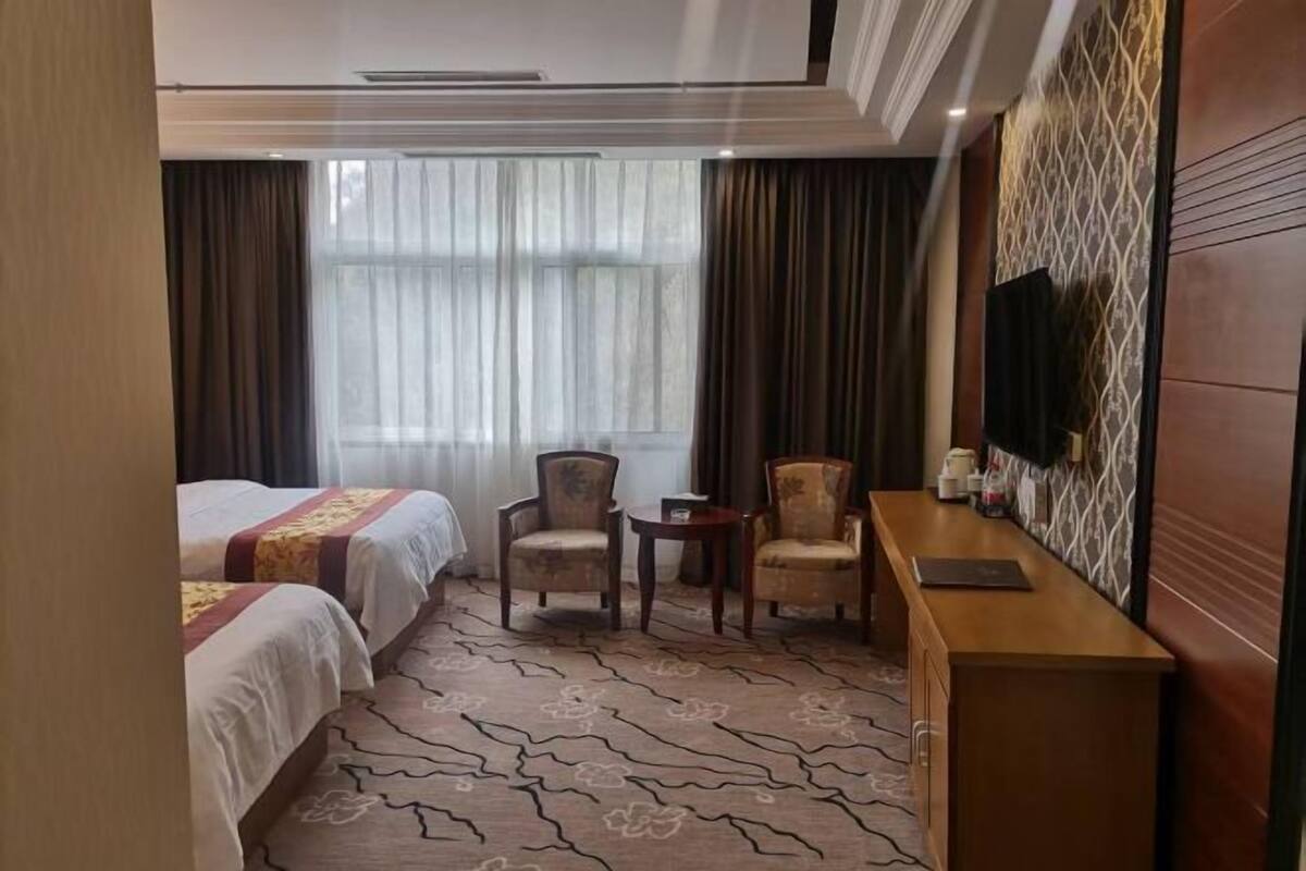 A hotel room with two beds, a small seating area with chairs, a wooden side table, a television, and a patterned carpet.