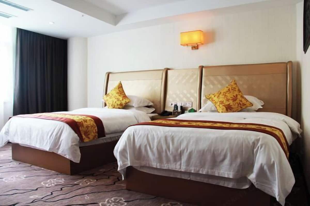 A hotel room with two beds, each with a headboard, white bedding, and decorative pillows. There is a wall-mounted lamp above the beds and a window with curtains.