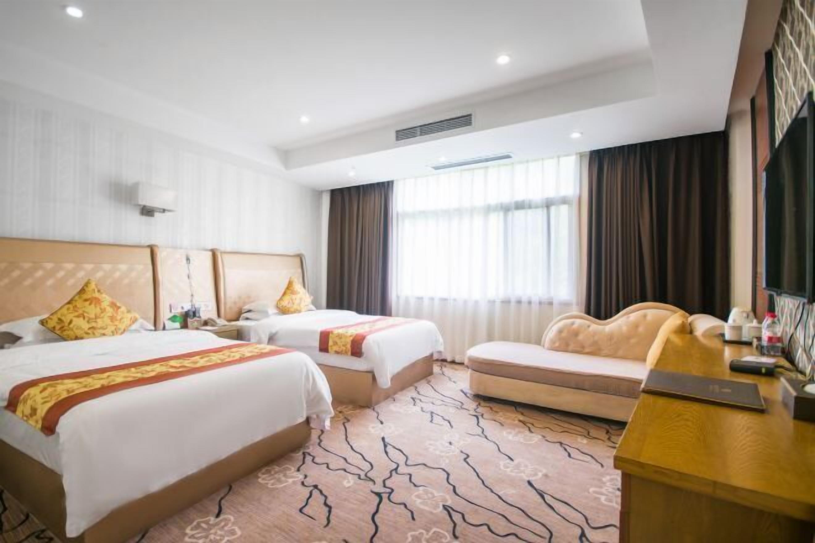 Hotellihuone, jossa on kaksi sänkyä, sohva, työpöytä ja suuri ikkuna, jossa on verhot.