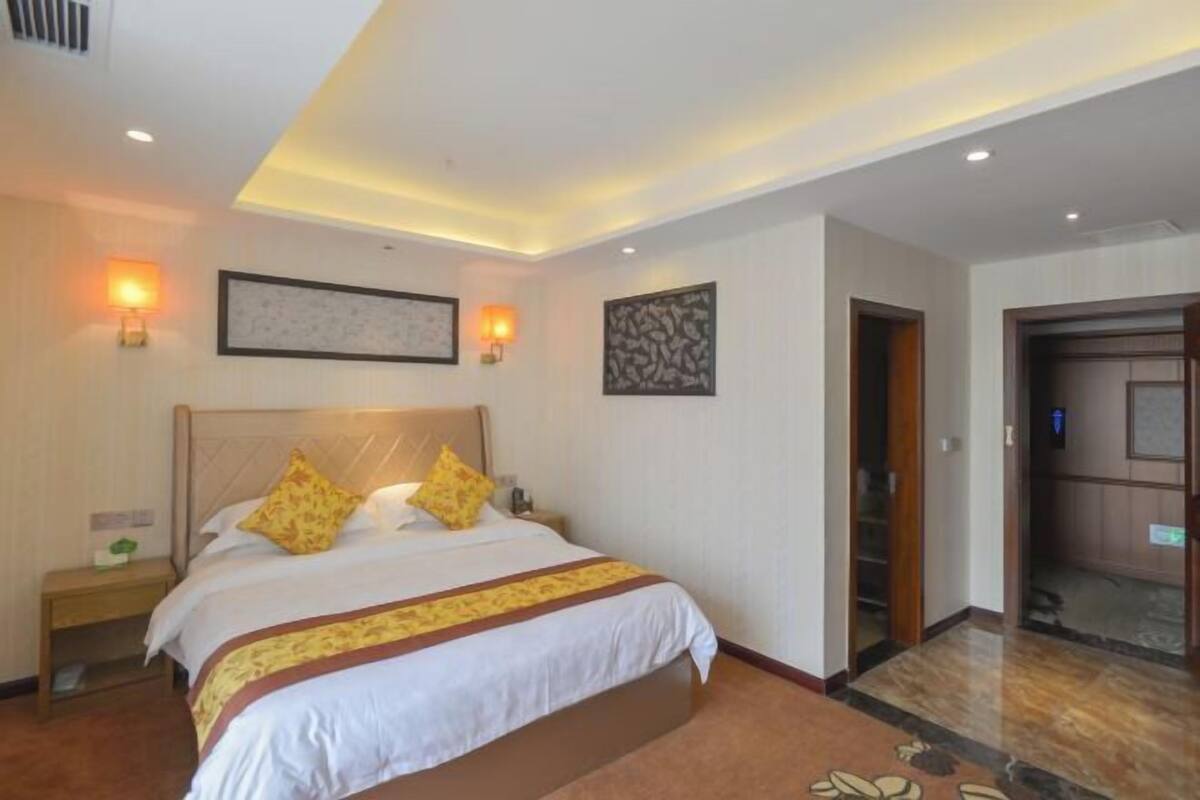 A hotel room with a large bed, a nightstand, a wall-mounted lamp, and a door leading to another room.