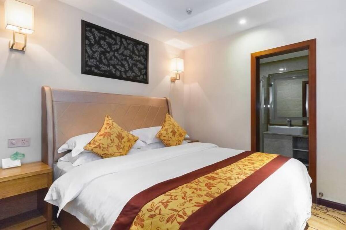 A hotel room with a large bed, two wall-mounted lamps, a framed artwork, and a bathroom visible through a glass door.