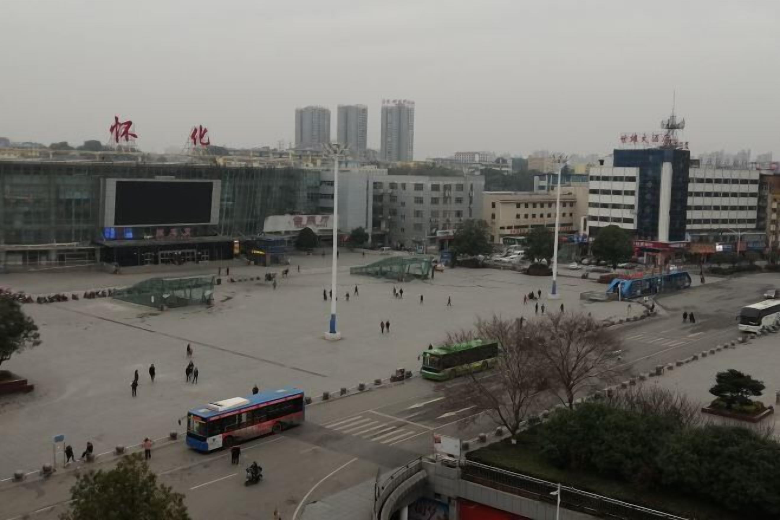 A spacious urban square with a large digital screen, modern buildings, and a bus in the foreground.