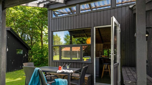 A covered outdoor patio with a table and chairs, a wooden deck, and a view of a house through large windows. - 4 Star Holiday Home in Hampen (Hampen)