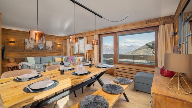A modern dining area with a wooden table set for six, a sofa, and a view of mountains.