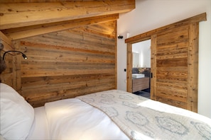 A bedroom with a wooden ceiling and walls, a bed with white bedding, and a bathroom visible through an open door.