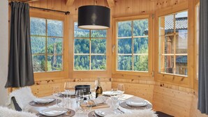A wooden cabin with a dining table set for six, large windows with a forest view, and a hanging light fixture.