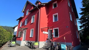 Un edificio rojo con múltiples ventanas y una puerta visible. Hay bicicletas estacionadas al frente y un coche de juguete pequeño en primer plano.