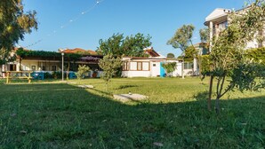 A grassy area with a small building, a blue door, and a white building in the background. - Korfez Pansiyon (Ayvalik)