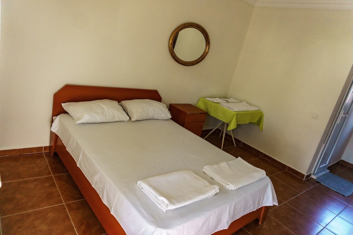 A neatly made bed with white linens and towels, a wooden headboard, a small bedside table with a green cloth, and a round mirror on the wall.