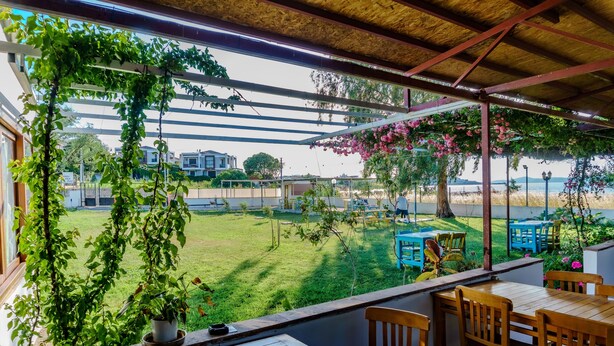 A covered outdoor seating area with wooden tables and chairs, a view of a grassy yard, and a pergola with climbing plants. - Korfez Pansiyon (Ayvalik)
