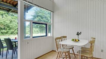 A room with a dining table and chairs, a window with a view of greenery, and a door leading outside.