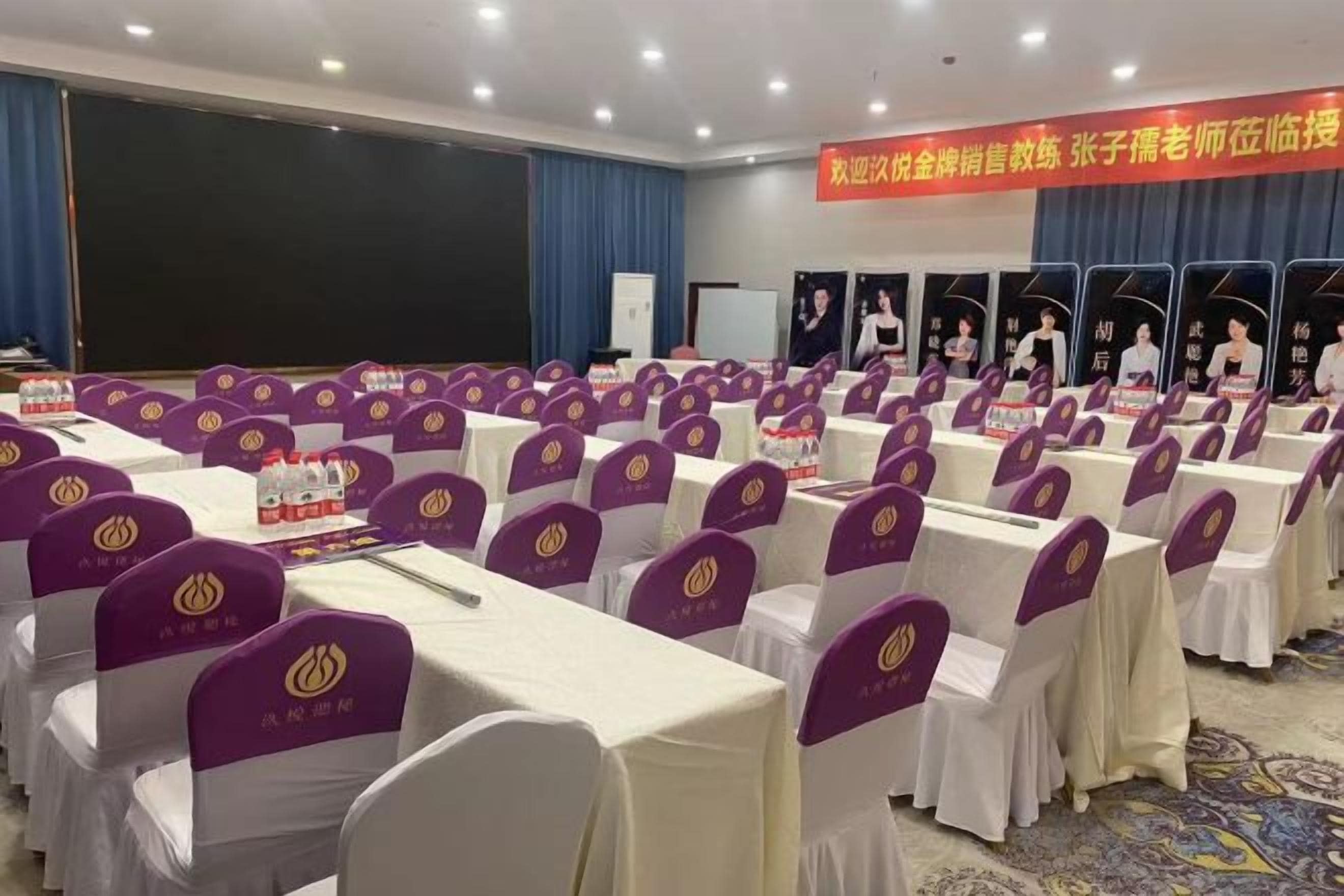 Un salón de conferencias con filas de mesas y sillas, una pantalla de proyector y un cartel con texto.