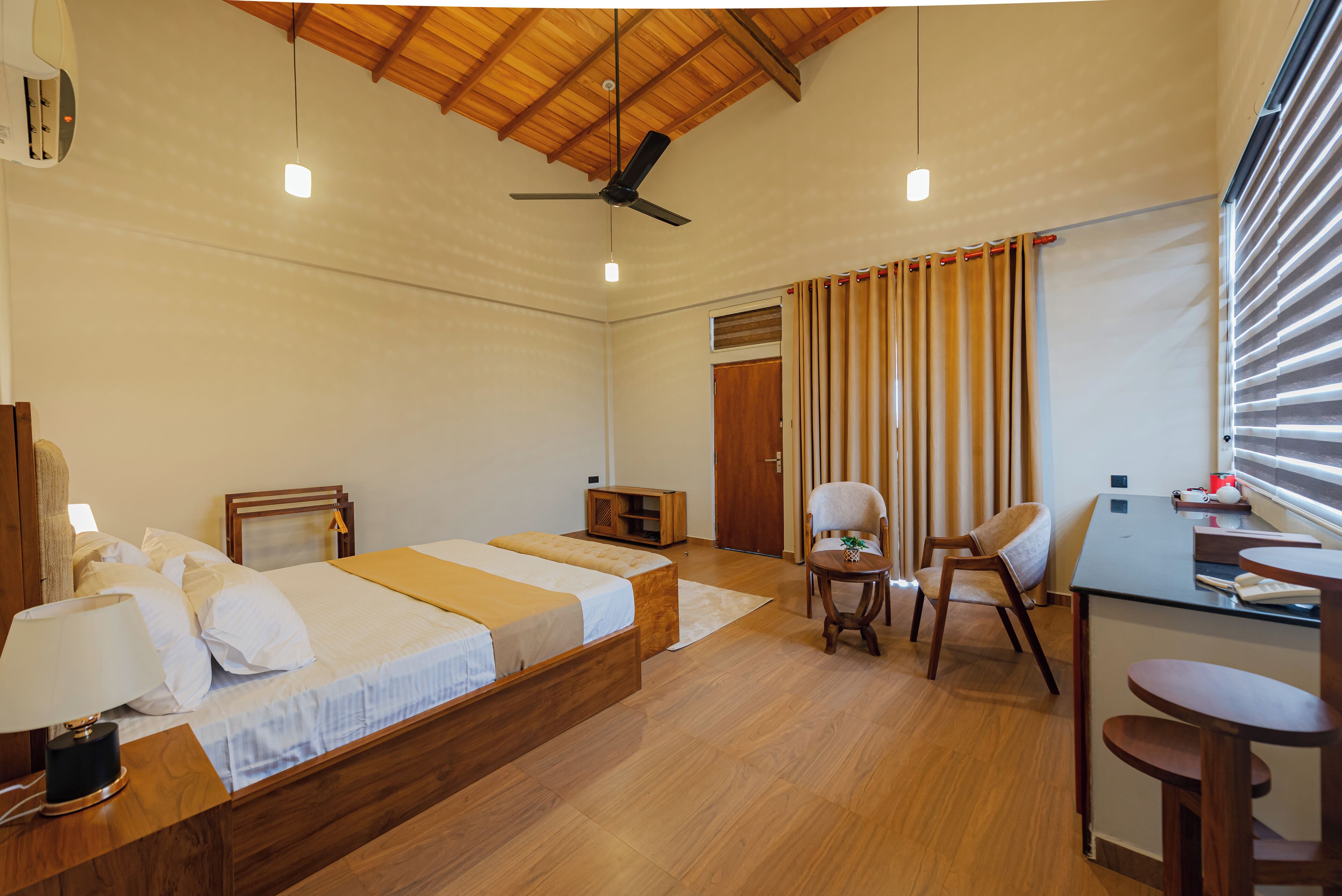 A hotel room with a wooden ceiling, a bed with white bedding, a desk with a chair, and a window with blinds.