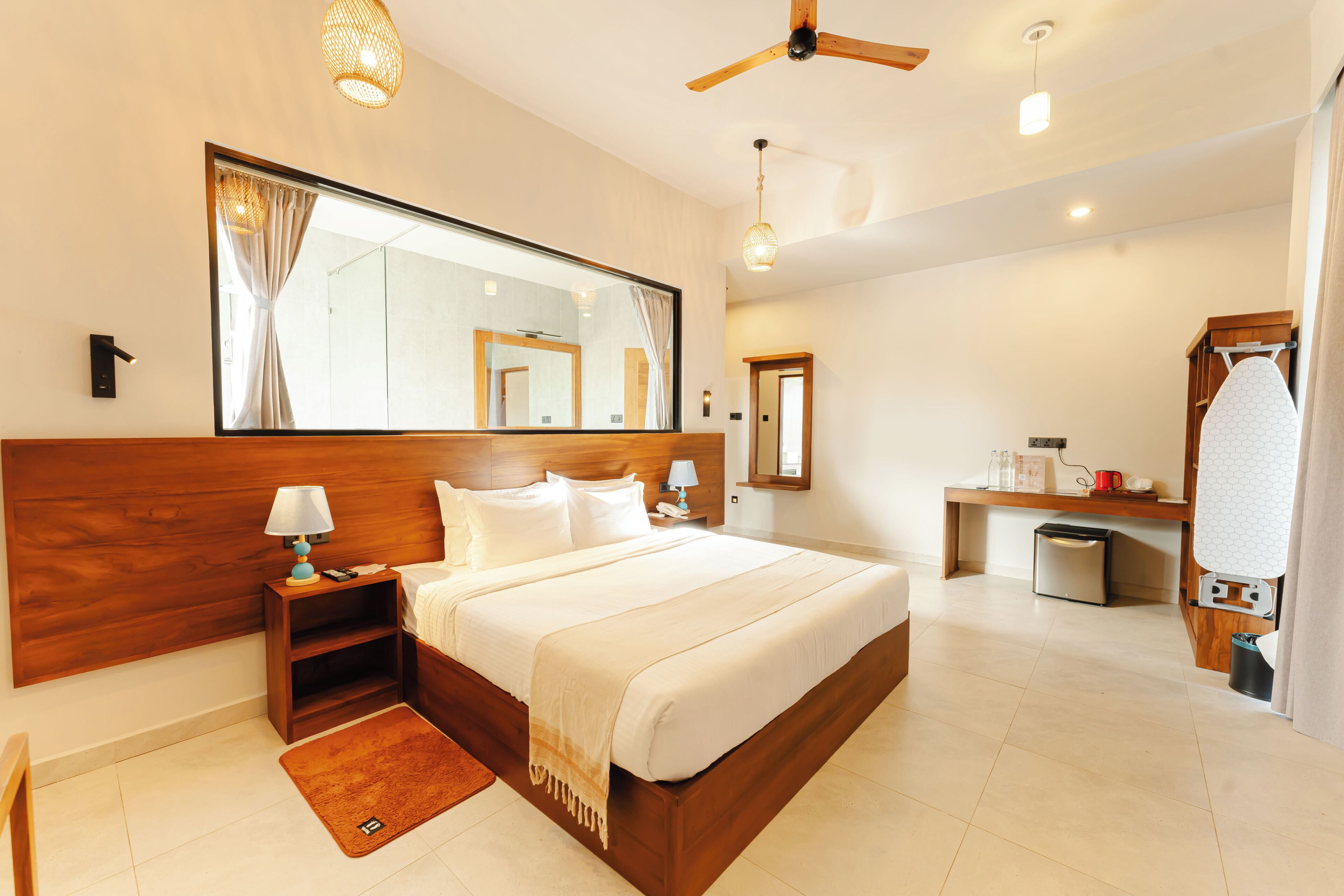 Une chambre d’hôtel avec un grand lit, une tête de lit en bois, des tables de chevet avec des lampes, un miroir, un ventilateur de plafond, un petit bureau avec une chaise et une salle de bain avec douche et toilettes.