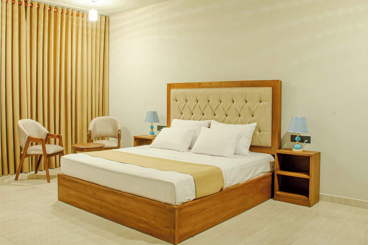 Un dormitorio ordenado con una cama de madera, dos sillas, mesitas de noche con lámparas y un telón de fondo de cortina.
