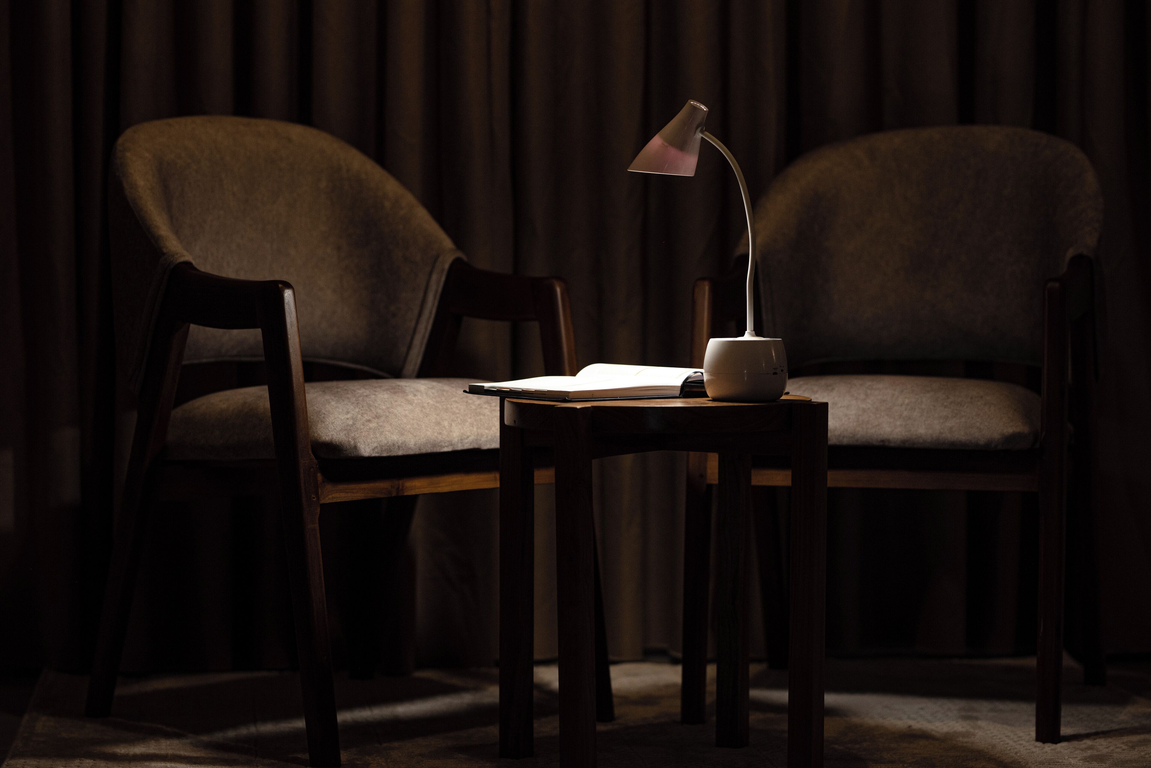 Una habitación oscura con dos sillas de madera y una pequeña mesa con una lámpara y un libro.