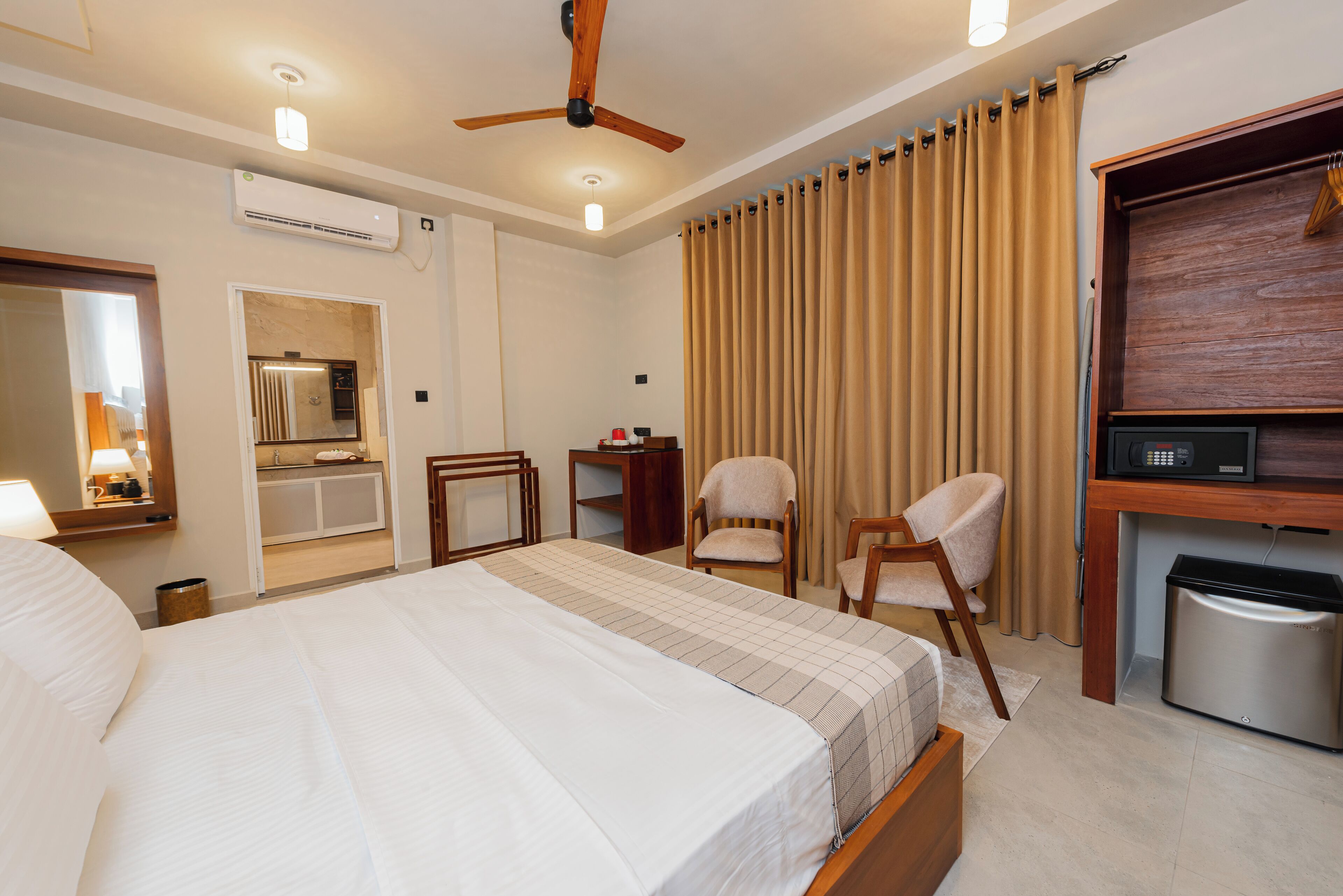 A hotel room with a large bed, a ceiling fan, an air conditioning unit, a desk with a microwave, a chair, and a small trash bin.