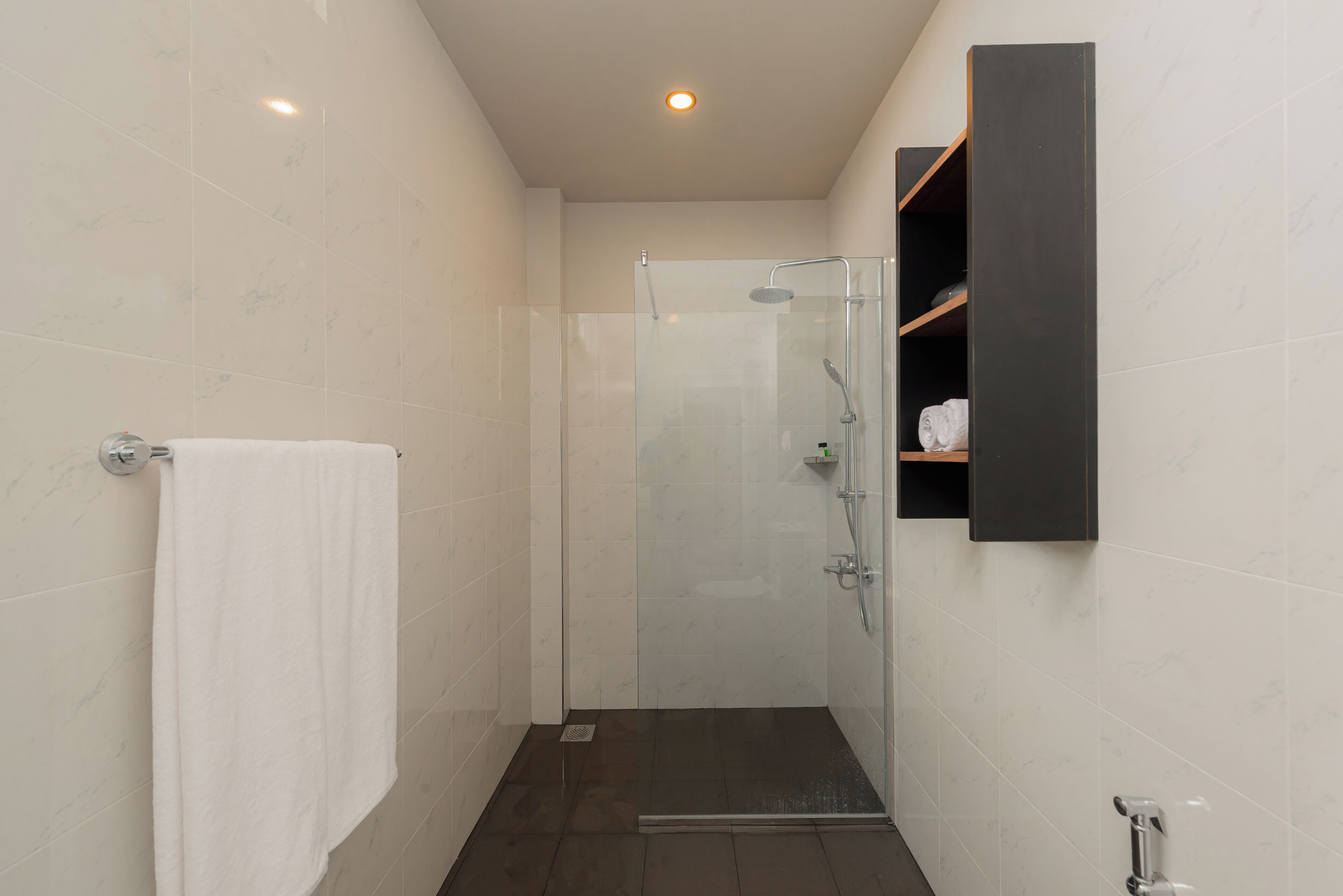 A modern bathroom with a glass-enclosed shower, a white towel hanging on a rack, and a dark wooden shelf with toiletries.
