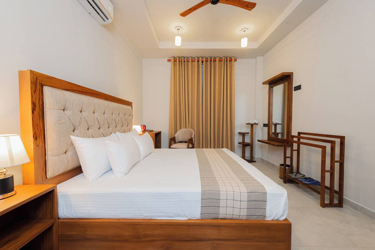 Un dormitorio ordenado con una cama grande, una mesita de noche con lámpara, una silla, un banco de madera y un espejo en la pared.