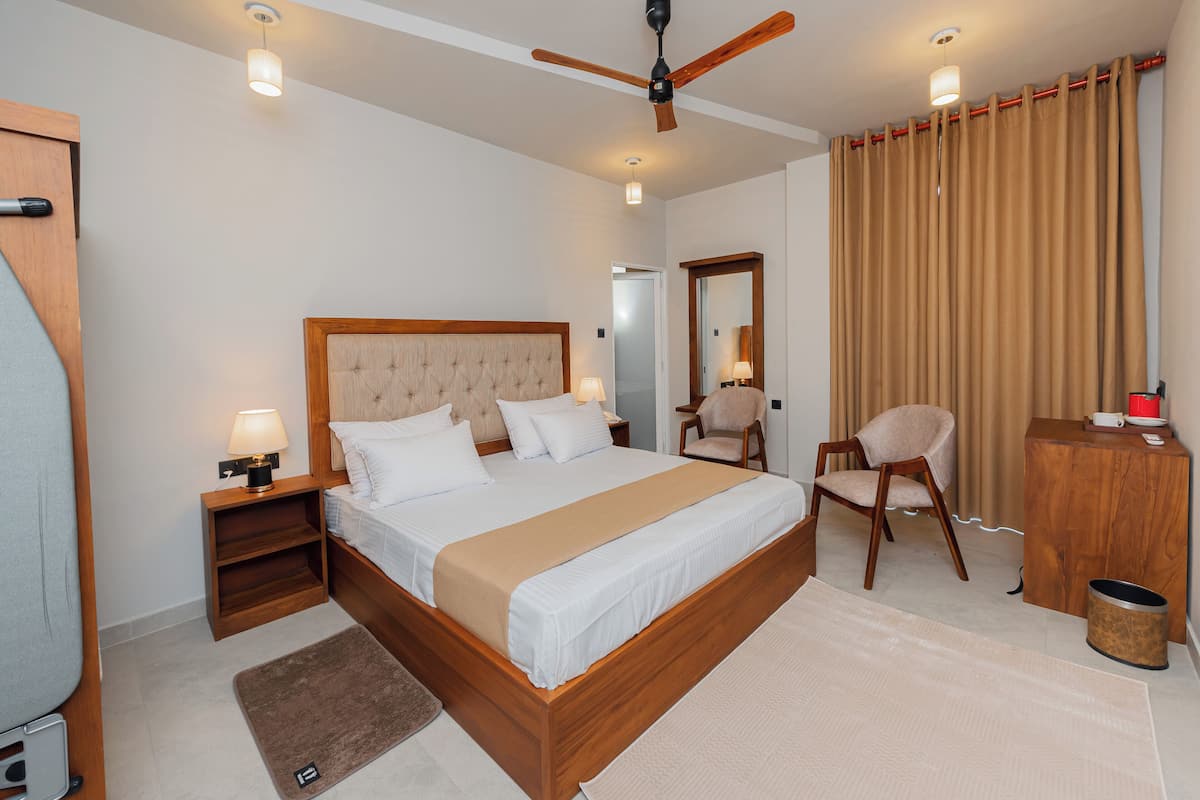 Una habitación de hotel moderna con una cama grande, cabecero de madera, lámparas de noche, ventilador de techo, una silla, un escritorio y un espejo.