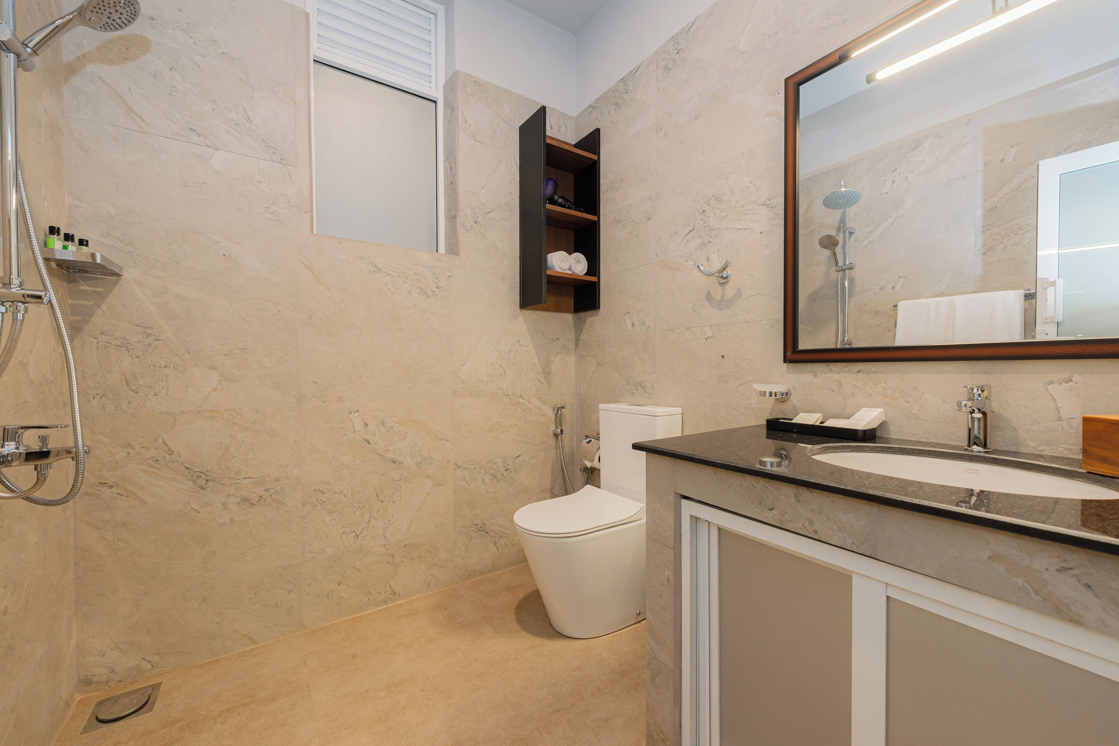 A bathroom with a marble floor, a white toilet, a shower with a handheld showerhead, a sink with a mirror above it, and a shelf with toiletries.