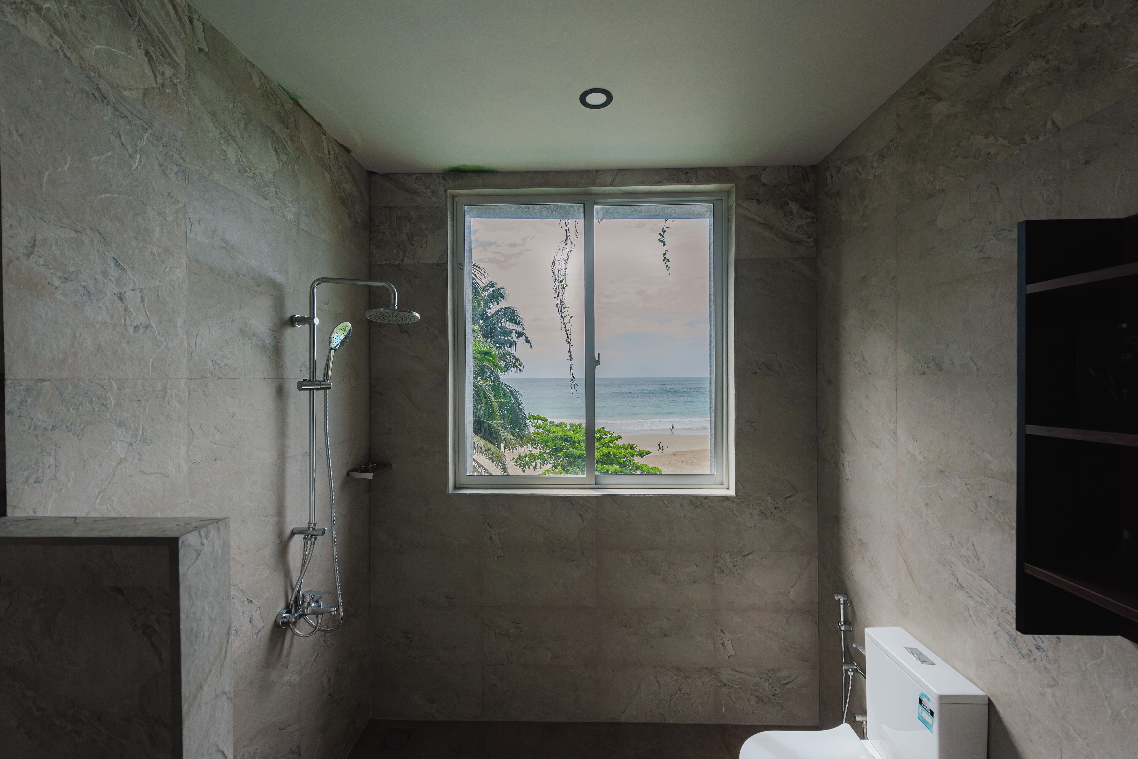 A bathroom with a shower, a toilet, and a window with a view of the beach.