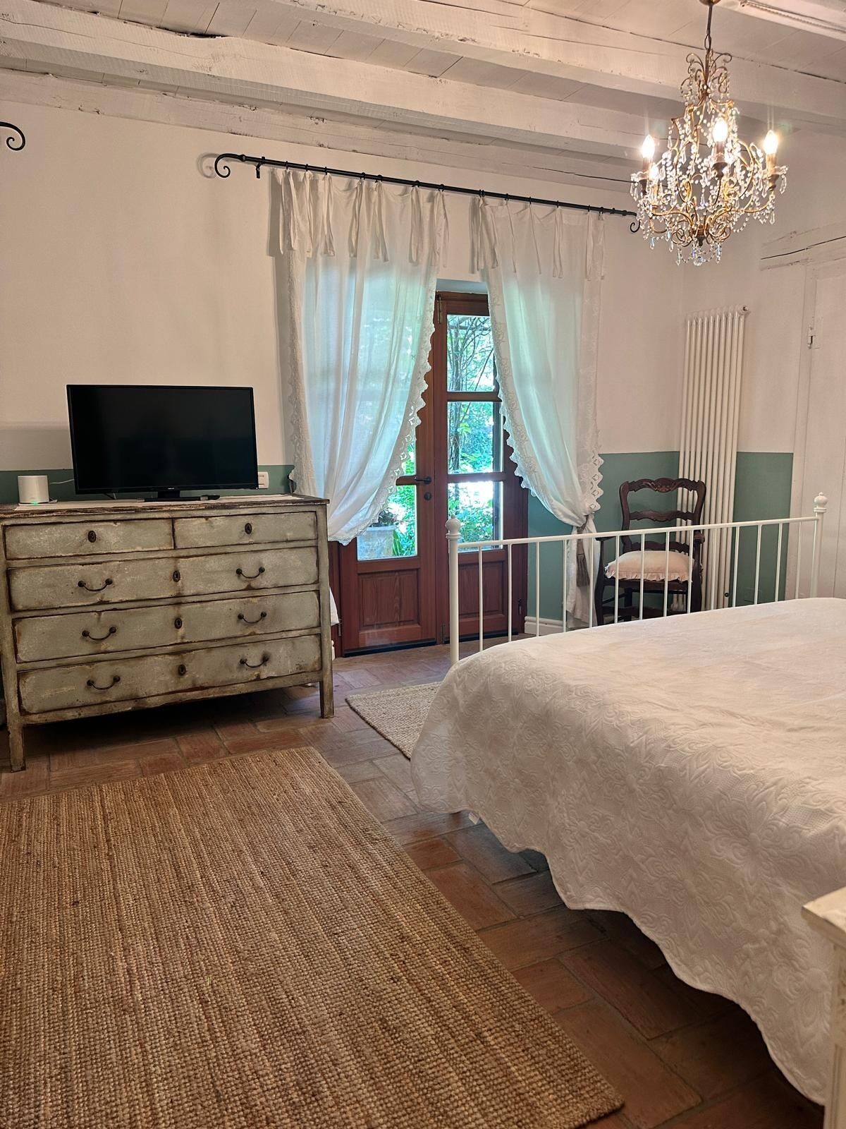 Una camera da letto con un letto, una cassettiera, una televisione, un lampadario e vista all'esterno tramite una porta.