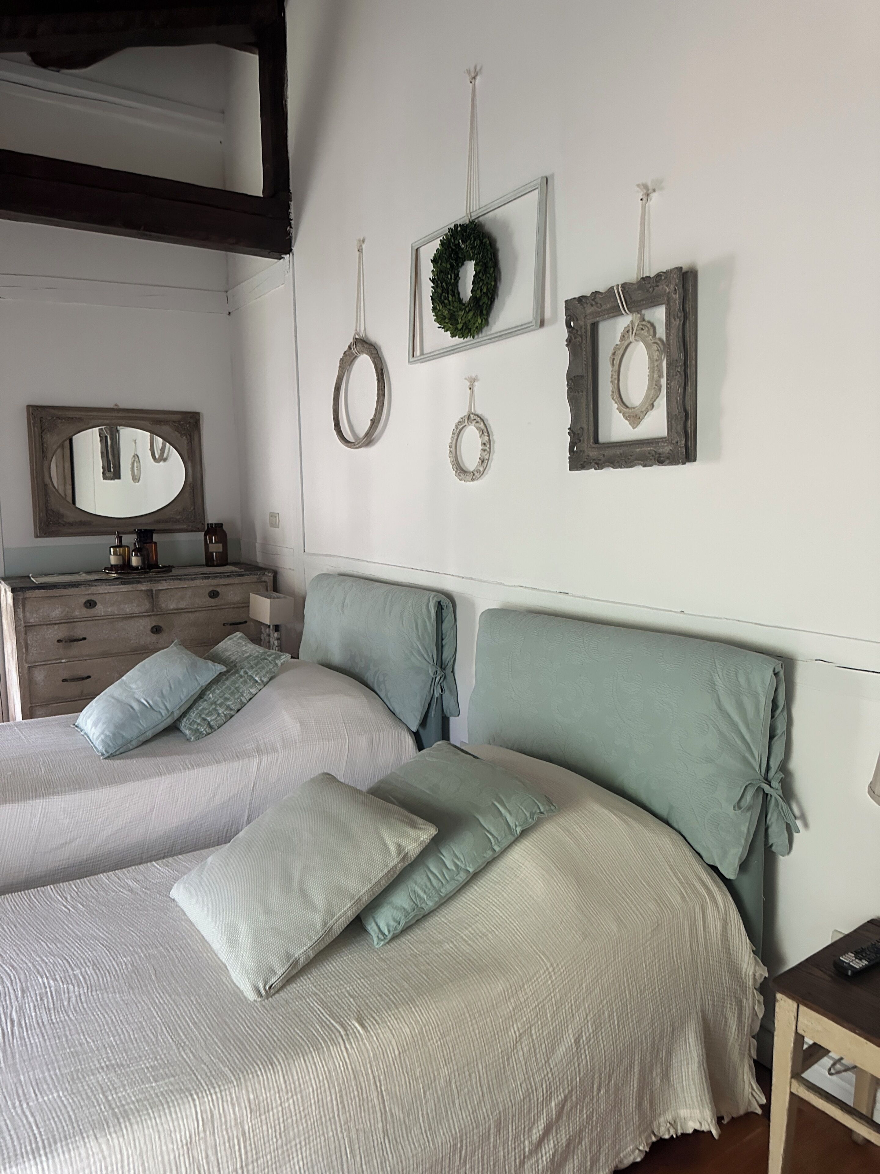 Ein Schlafzimmer mit zwei Betten, einem Schminktisch und Wanddekorationen, darunter ein Spiegel, ein Kranz und gerahmte Bilder.