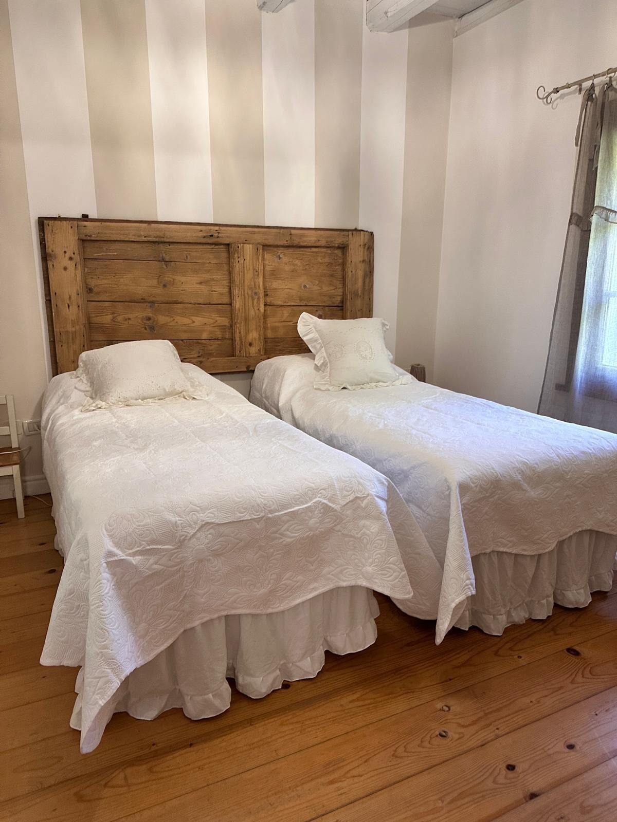 Ein Zimmer mit zwei Einzelbetten, einem hölzernen Kopfteil, weißer Bettwäsche und einem Fenster mit Vorhängen.