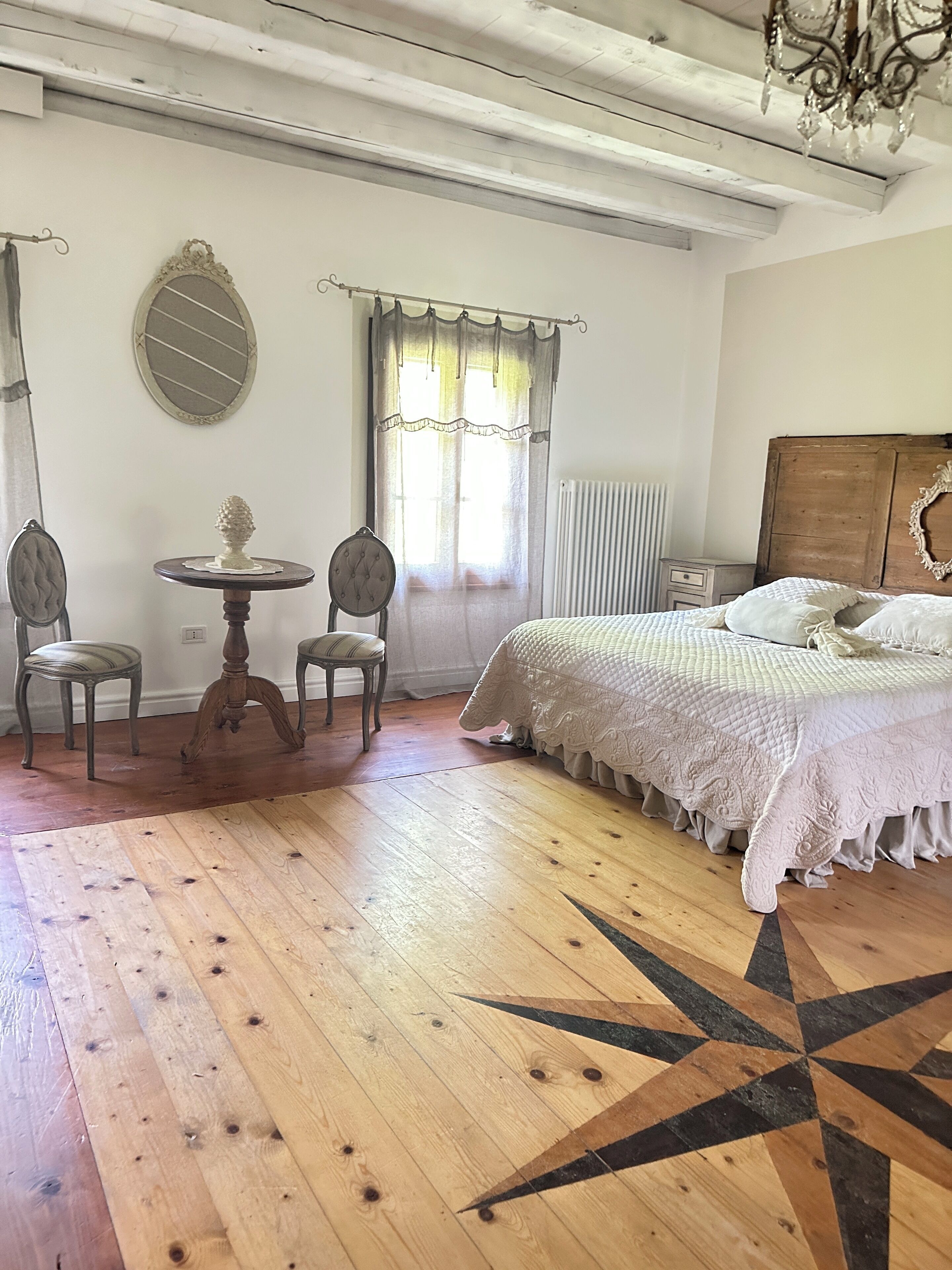 Un dormitorio con piso de madera, una cama con colcha blanca, dos sillas, una mesita, un espejo y un ventanal con cortinas.