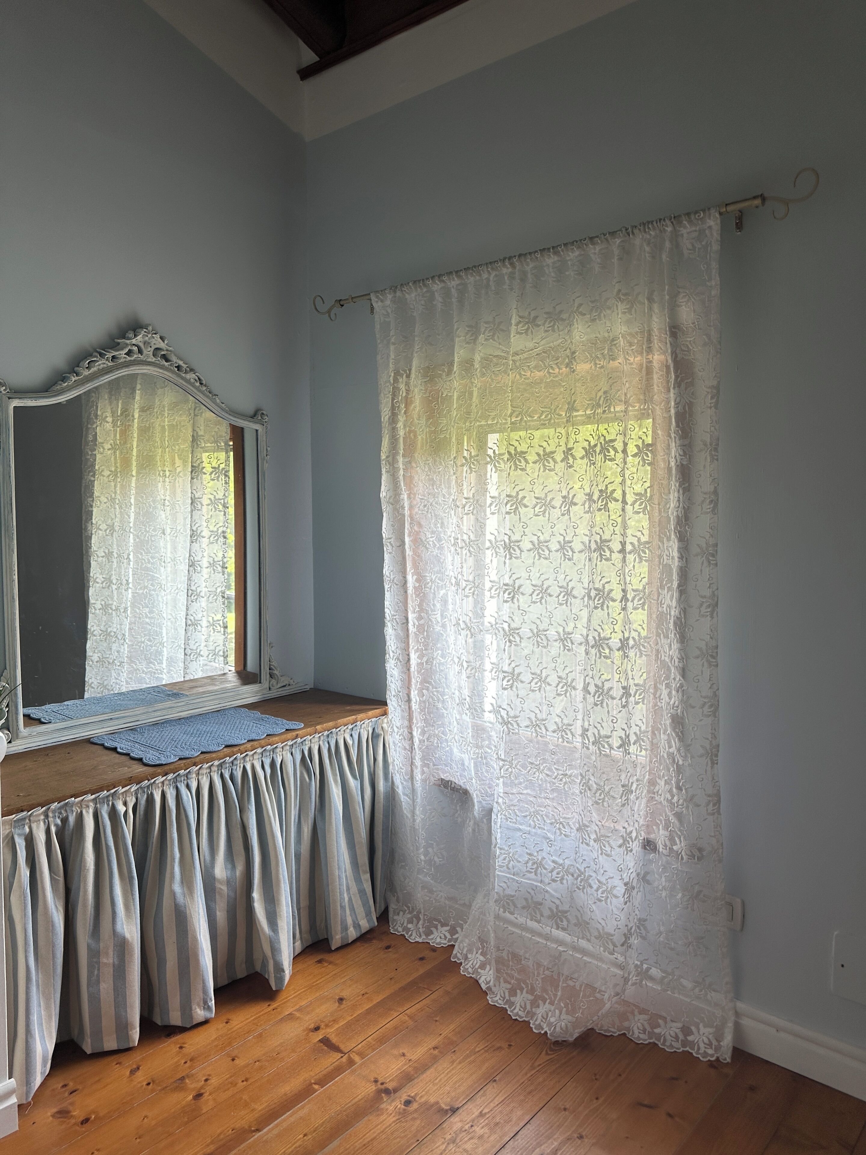 A room with a wooden floor, a mirror with an ornate frame, a wooden bench, and a window with a sheer white curtain.