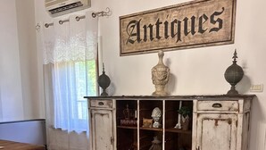 A room with a wooden floor, a white cabinet labeled "Antiques" with decorative items, a window with white curtains, and an air conditioning unit. - BednBarchessa (Portogruaro)