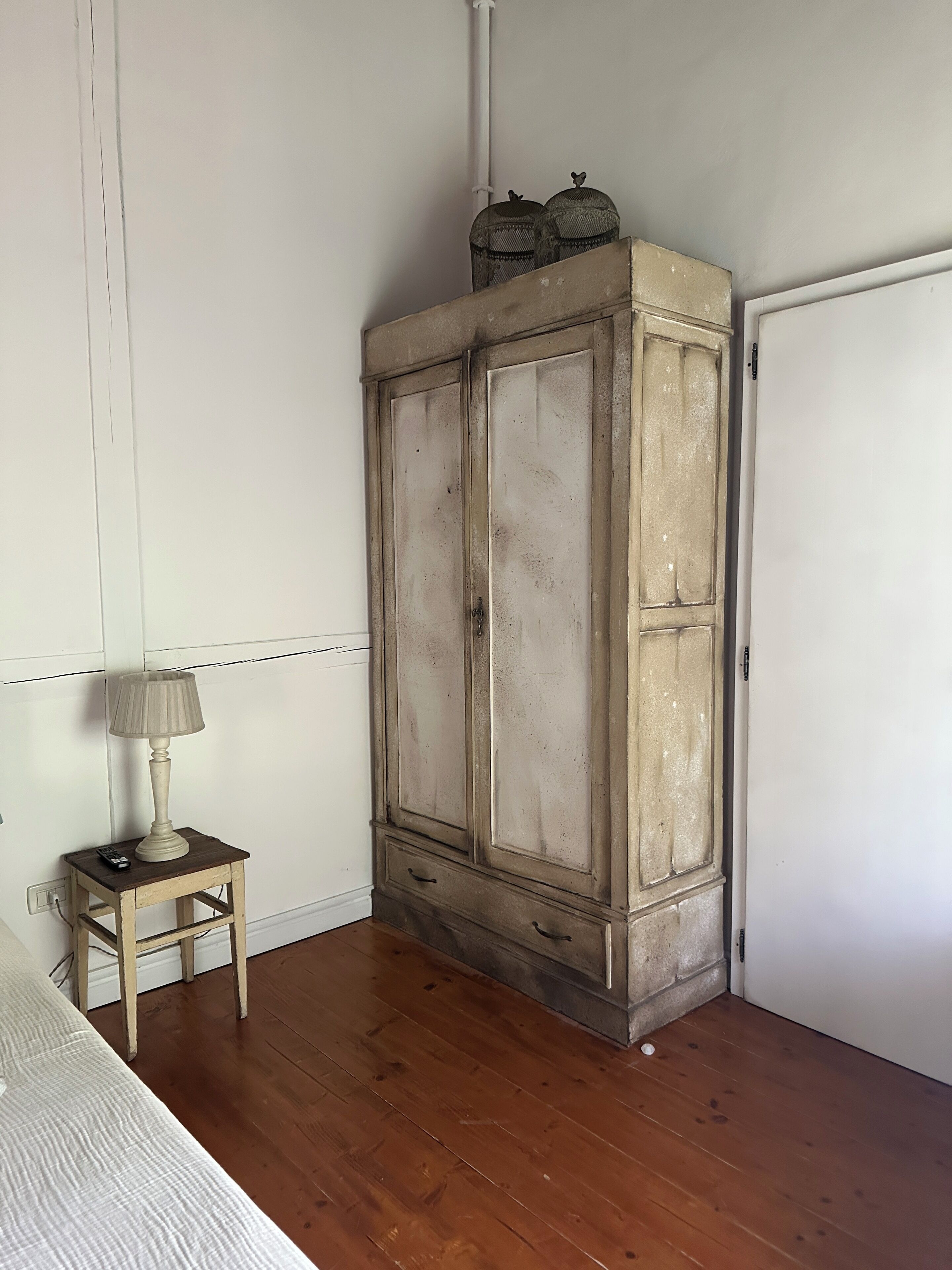 Une pièce avec un plancher en bois, une petite table en bois sur laquelle se trouve une lampe, et une armoire blanche surmontée d’une cage à oiseaux en métal.