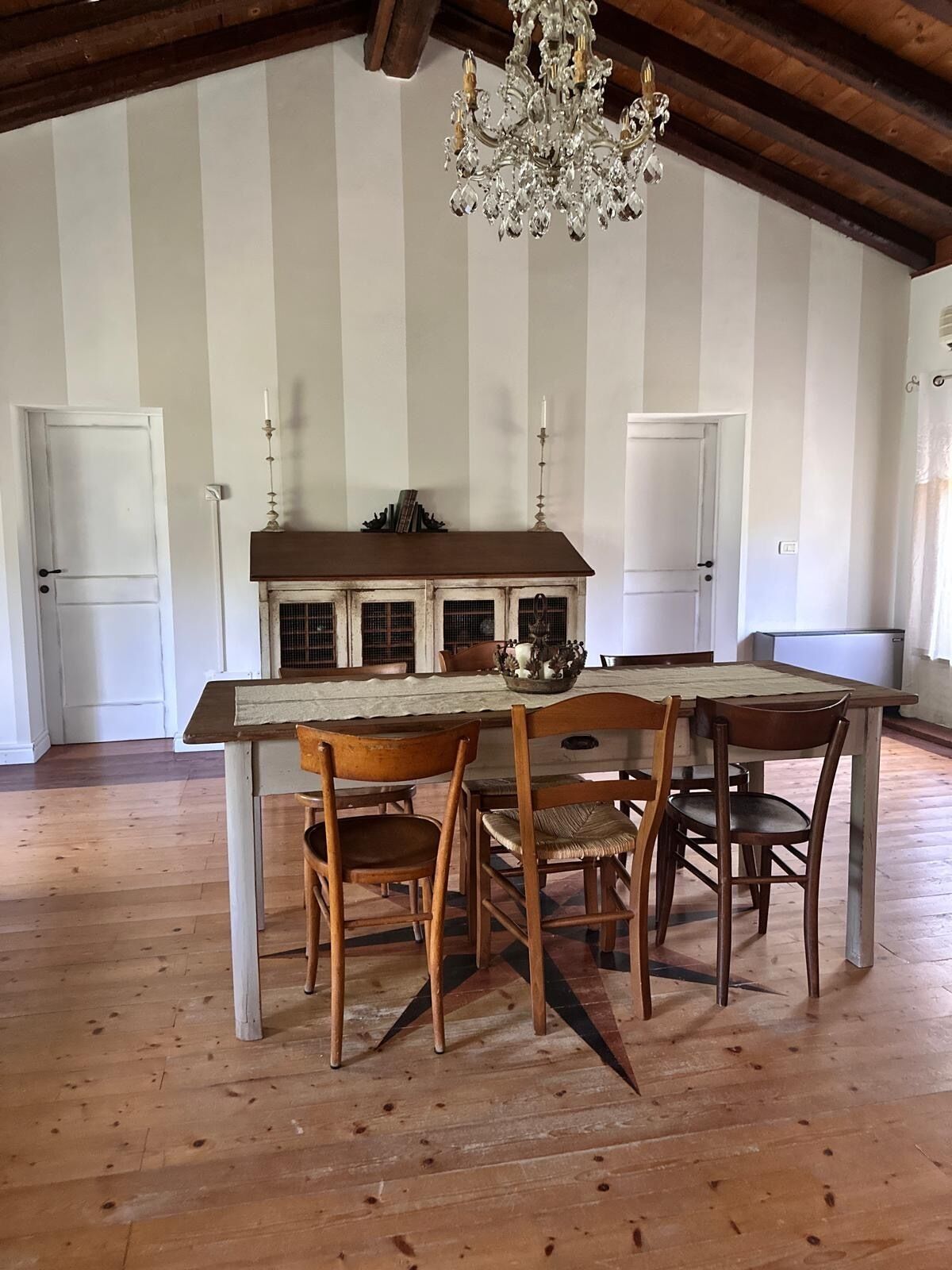 Uma área de jantar rústica com mesa e cadeiras de madeira, um candelabro e uma parede com listras brancas.