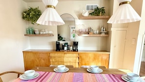 Un coin repas avec une table en bois dressée pour six personnes, un chemin de table à rayures et, au fond, une cuisine avec des étagères et une plante.