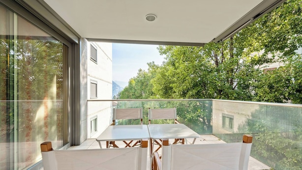 A balcony with a table and chairs, large windows, and a view of trees and buildings.