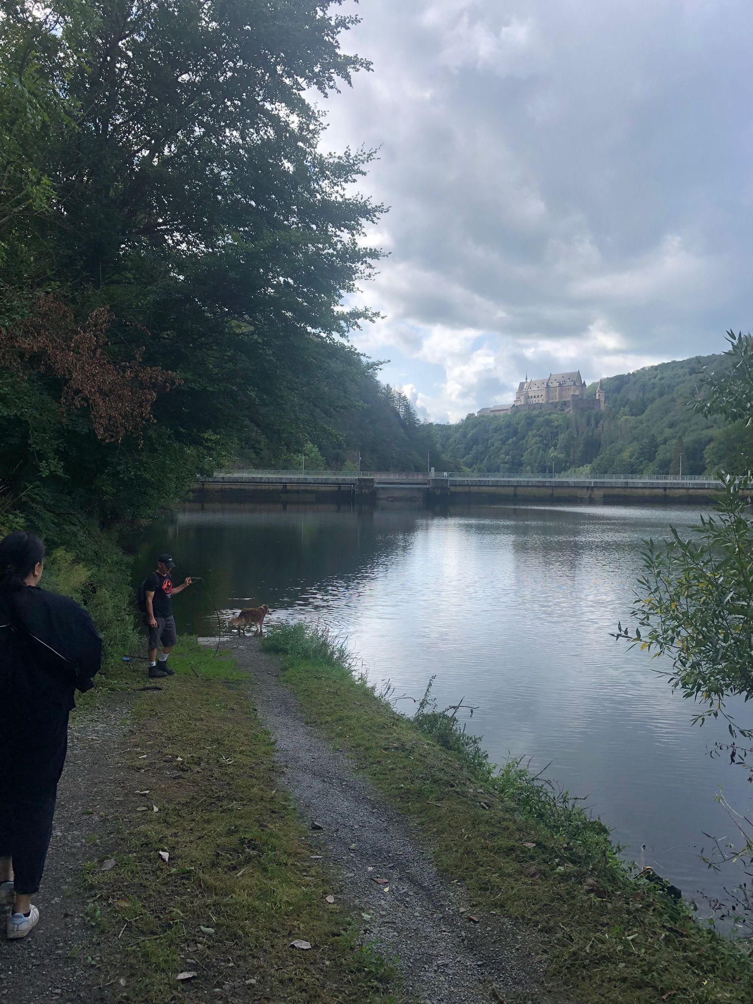 A calm river with a bridge, lush greenery, and a castle on a hill in the background.