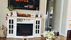 A living room with a fireplace, a flat-screen TV displaying Netflix, a tray table with drinks and snacks, and a door leading to another room. - Luxury Home w/private Pool, Mt Views, 85” Roku Movie Room, Arcade, & King Bed! (Talbott)