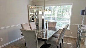 Un coin repas comprenant une table à plateau en verre, quatre chaises, un lustre et une fenêtre avec des stores.